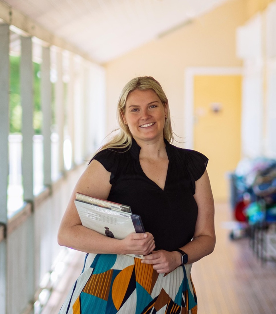 Teacher holding workbooks, near bag racks on a verandah of a school building.