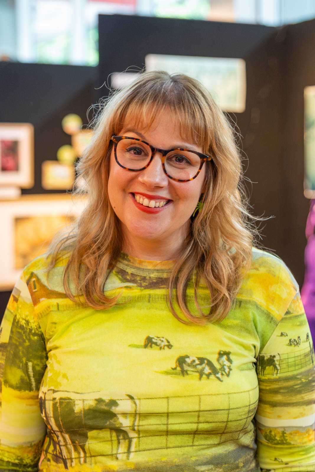 Alice is wearing glasses and a green-yellow printed top in front of an art exhibition.